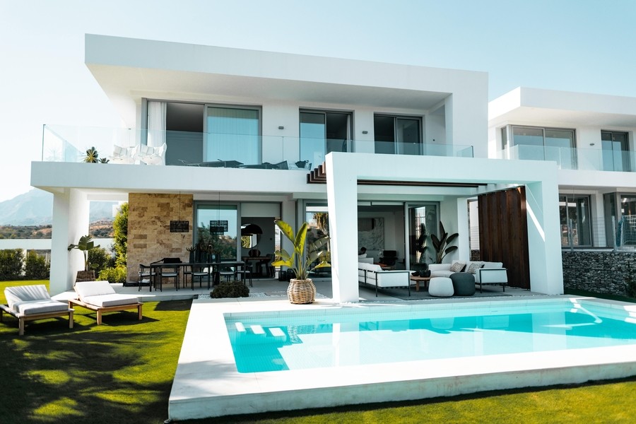 A modern, white home with an expansive patio, a pool in the foreground, as well as green grass and lawn chairs.