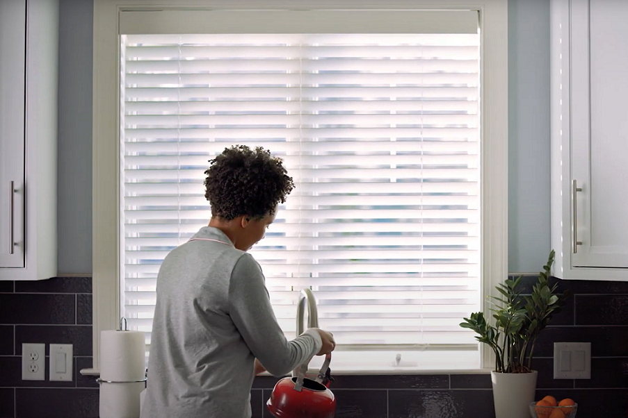 A large kitchen window with motorized blinds, and a woman at the kitchen sink.