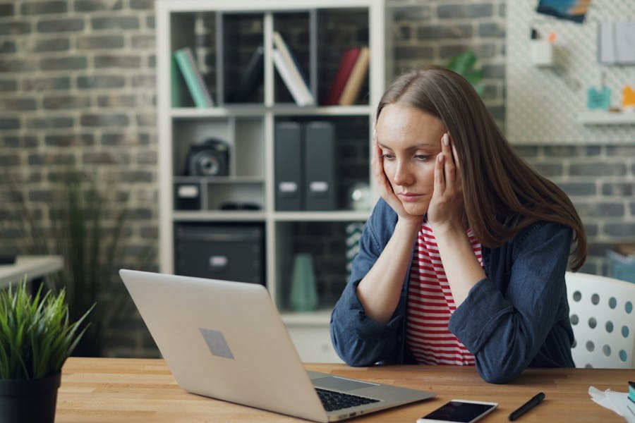 Frustrated young woman sitting at a desk in front of a laptop, resting her head in her hands, with a smartphone and potted plant on the desk in a home office setting.