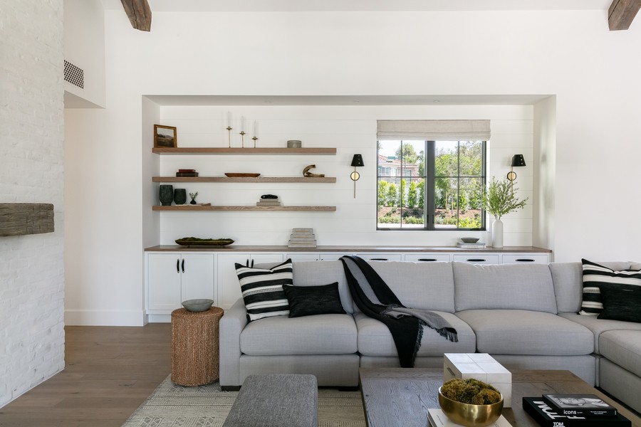 A modern living room with a light gray sectional sofa, black and white accent pillows, built-in white cabinetry with open wood shelves styled with minimal decor, a large window with a Roman shade, and neutral-toned accessories.