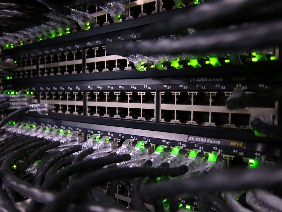 A view of the back of a networking equipment rack with many cables and green lights.